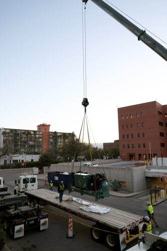 An industrial chiller suspended in mid-air by a crane, surrounded by workers in safety gear. The chiller, a large metallic structure, dangles from heavy-duty straps as it is carefully maneuvered into place for installation on this retrofit, government HVAC project.