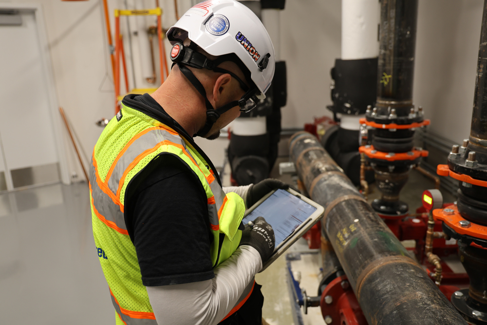 ACCO Engineered Systems technician performing mechanical system commissioning and quality assurance in a commercial building using a tablet.