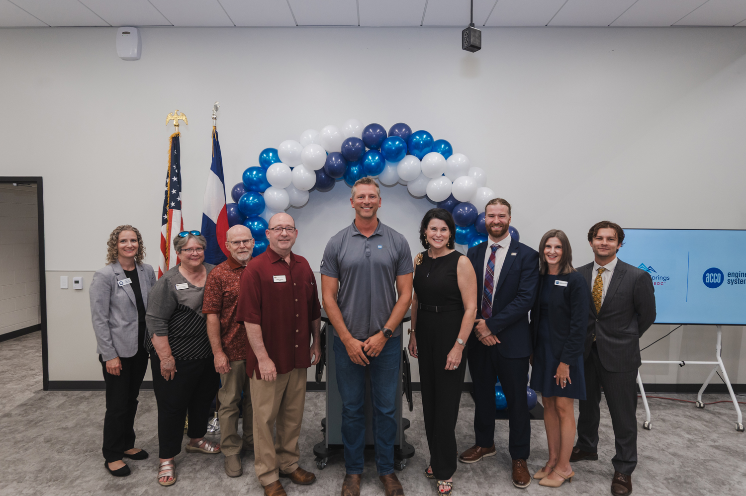 A group of ACCO Engineered Systems representatives, local elected officials and community partners pose for a photo during the grand opening celebration of ACCO's new fabrication facility in Monument, Colorado. Blue and white balloon arches, U.S. and Colorado flags, and an ACCO display screen are visible in the background.